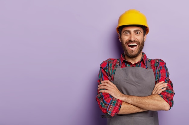 Positive happy foreman in workwear, keeps arms folded, wears yellow hardhat