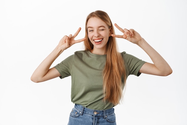 Free photo positive and happy young blond girl, winking and smiling white teeth, showing peace v-sign gesture near eyes, standing joyful against white wall