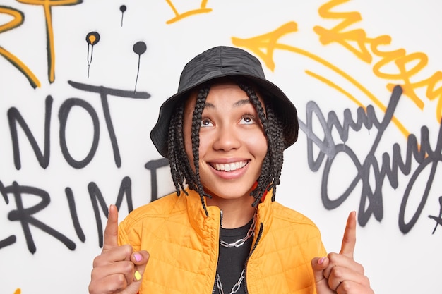 Free photo positive hipster girl with braids smiles broadly points index fingers overhead dressed in fashionable clothes demonstrates something against graffiti wall belongs to youth subculture