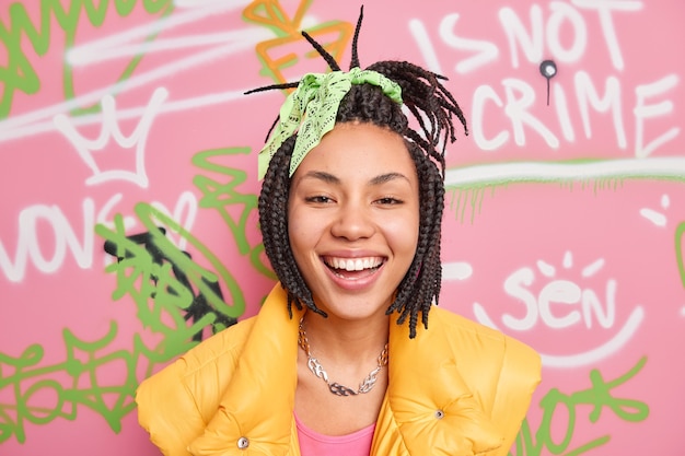 Free photo positive hipster gurl smiles broadly enjoys spare time poses against colorful graffiti wall dressed in casual street style clothes being in good mood