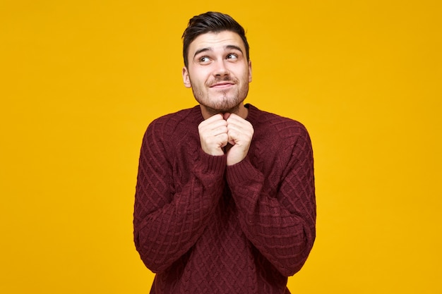 Positive human emotions, feelings and facial expression. Picture of handsome young European man with dark hair and stubble looking up with cute mysterious smile, holding hands at his face, dreaming