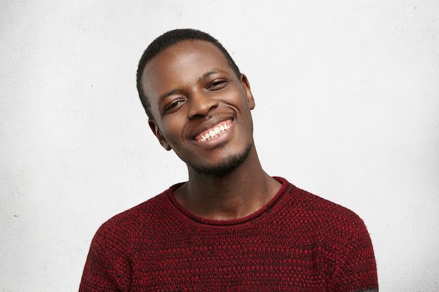 Positive human facial expressions and emotions. Headshot of handsome happy dark-skinned man dressed in casual sweater smiling kindly, showing his white teeth while pleased with compliment or good news