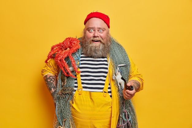 Positive impressed bearded sailor in striped vest poses with red octopus on shoulder, holds smoking pipe, carries fishing net