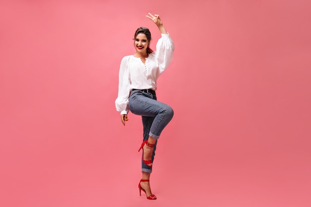 Free photo positive lady in denim outfit shows peace sign on pink background.  happy young woman in good mood in white blouse smiling..