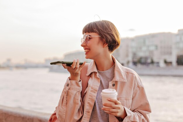 Positive lady holding phone on background of river Shorthaired woman in light jacket and glasses with backpack posing with cup of tea outside