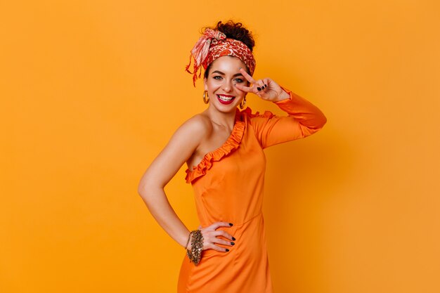 Positive lady in orange elegant dress and fashionable headband sincerely smiles and shows peace sign on orange space.