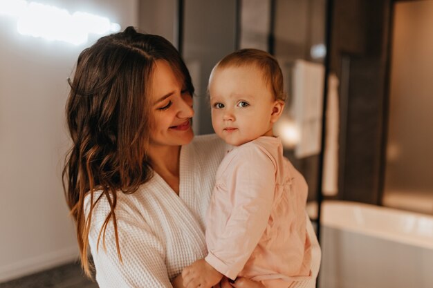 Positive long-haired woman in white robe with tender smile looks at her little daughter.