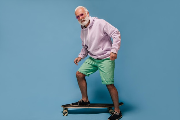 Free photo positive man in hoodie and sunglasses riding longboard modern guy with white beard in bright outfit posing on isolated background