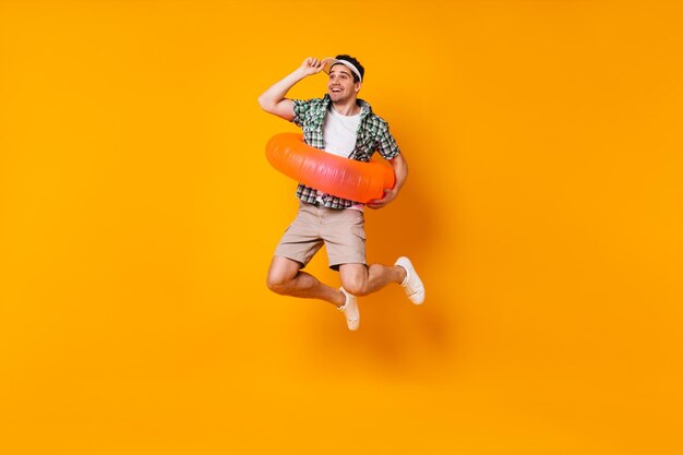 Positive man is jumping on orange background Portrait of guy in cap and summer outfit with inflatable circle