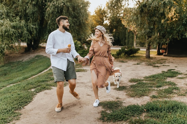 Positive man with ice cream is holding hand of smiling woman in brown dress Romantic couple walking with big labrador in park