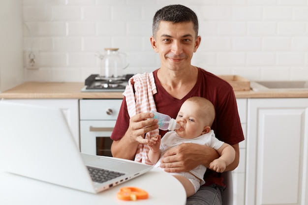 Positive man with smile wearing burgundy casual t shirt with towel on his shoulder, looking after baby and working online from home, giving water to his daughter, looks at camera.