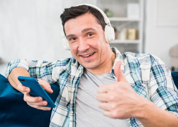 Free photo positive man with thumb up listening music in headphones and browsing on smartphone on sofa