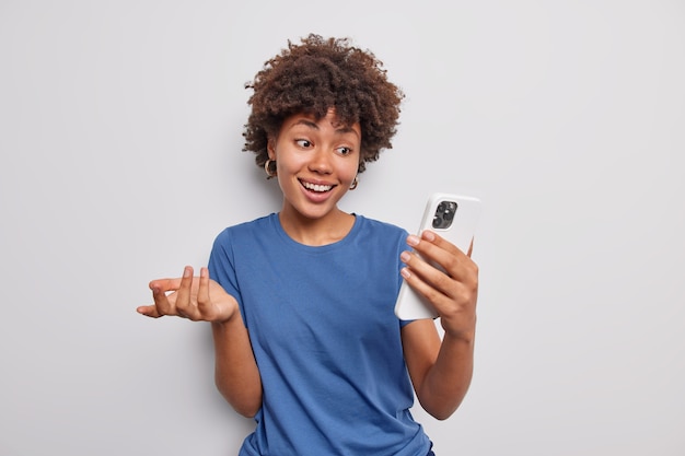 Positive millennial girl makes video call communicates with best friend distantly holds mobile phone uses free internet connection dressed in casual blue t shirt isolated over white background