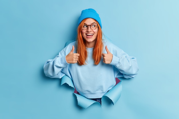 Free Photo positive millennial girl with natural ginger hair recommends sales keeps thumbs up and smiles gladfully gives approval wears blue hat and sweatshirt breaks through paper hole