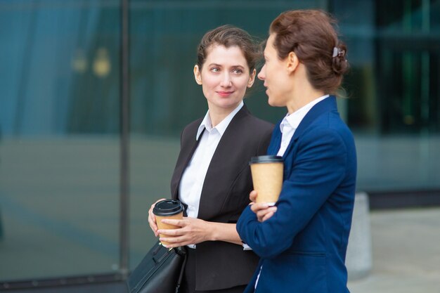 Positive office female friends with takeaway coffee cups walking together outdoors, talking, discussing project or chatting. Medium shot. Work break concept