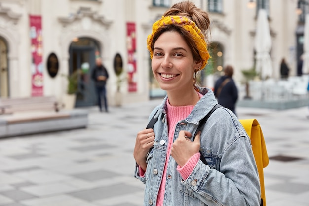 Positive optimistic woman has happy expression, wears stylish clothes, has yellow small rucksack on back, wanders across city streets