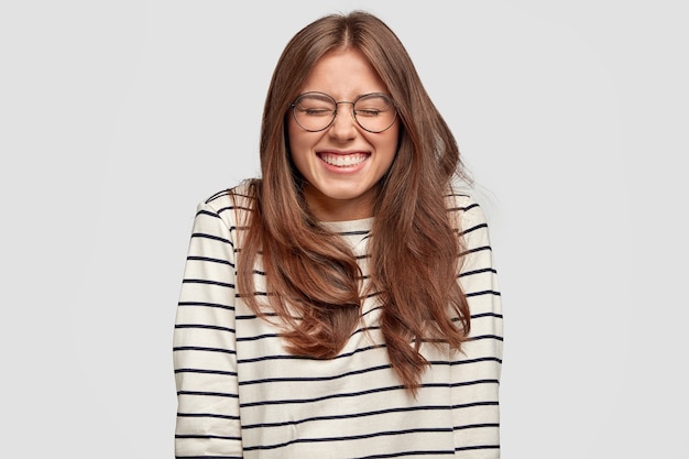 Free photo positive optimistic young female smiles joyfully, has white even teeth, dressed casually, being in high spirit, expresses happiness, spends free time with friends, isolated over white wall