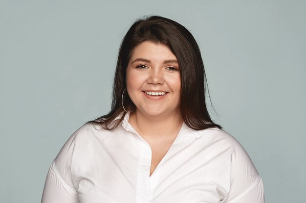 Positive reaction, attitude and life perception. Cute charming young Caucasian woman with curvy body posing at gray office wall and smiling cheerfully, rejoicing at her career success