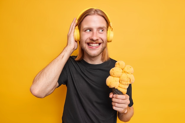 Free photo positive redhead bearded man keeps hand on headphones holds delicious ice cream enjoys favorite music via headphones dressed casually isolated over yellow wall. hipster guy with gelato