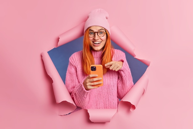 Positive redhead woman points at display of modern smartphone gets useful information from intenet surfs in social networks giggles positively uses mobile app or new technology. Blogger lifestyle.