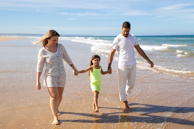 Positive relaxed family couple and little girl walking on wet golden sand on beach, kid holding parents hands