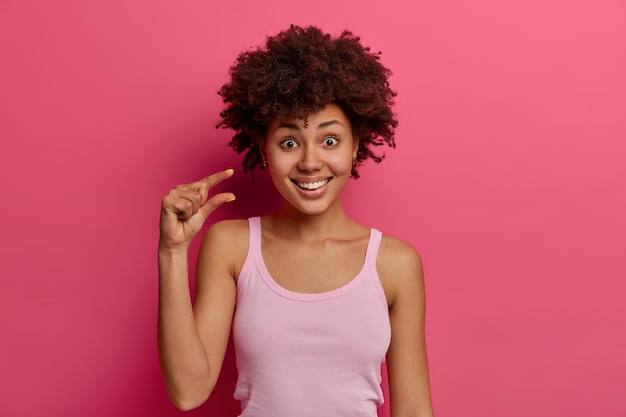 Positive satisfied dark skinned young woman makes small size sign, demonstrates something very little, gestures with hand and shapes object, wears casual vest, isolated over pink wall.