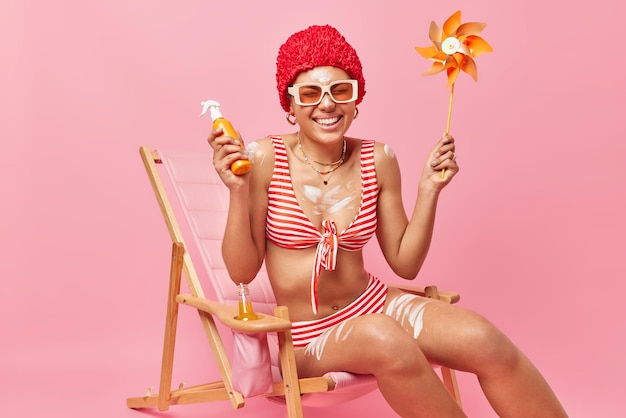 Free Photo positive slim woman dressed in swimwear has perfect summer holidays applies sunscreen lotion holds bottle and windmill poses on deck chair isolated over pink background skin protection from sunburn