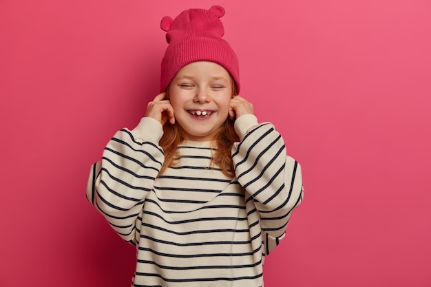 Free photo positive small child plugs ears, ignores loud sound, closes eyes and chuckles, wears pink hat and oversized striped jumper, poses indoor. little girl being naughty, doesnt want to hear parents