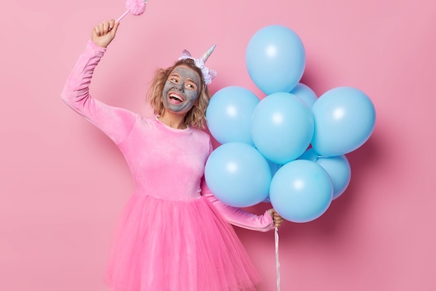 Free photo positive smiling carefree woman prepares for holiday applies clay nourishing mask on face holds bunch of blue helium balloons wears festive dress dances carefree isolated over pink background