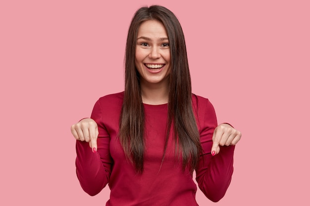 Positive smiling lady with long dark hair points with both fore fingers down, being in high spirit, recommends looking on floor