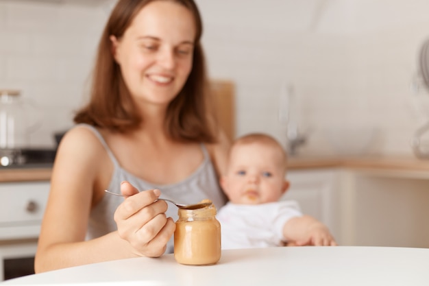 Positive smiling young adult dark haired mother feeding her little daughter with fruit or vegetable puree, holding spoon with healthy food, posing in kitchen.