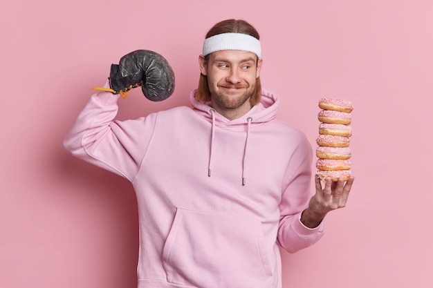 Free Photo positive sportsman raises arm shows biceps after training wears boxing glove holds pile of delicious doughnuts dressed in sweatshirt headband.