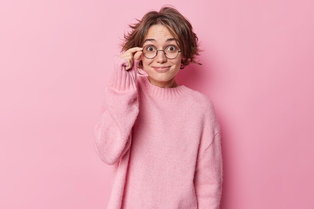 Positive surprised woman keeps hand on rim of spectacles smiles pleasantly reacts on good news wears casual cashmere jumper hears something awesome isolated over pink background. Reaction and emotions