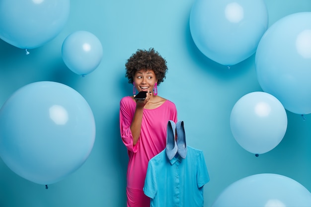 Free Photo positive talkative woman makes voice call, consults with friend what better to wear for theme party, holds blue shirt and shoes, dressed in pink dress, poses indoor against big helium balloons