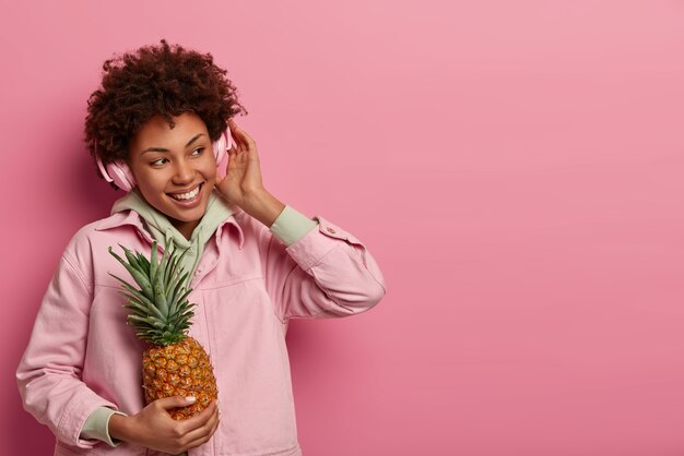 Positive teenage girl listens music, wears headphones on ears, feels very glad, holds ripe pineapple, looks aside with happiness, wears casual clothing, isolated over rosy wall, blank space for promo