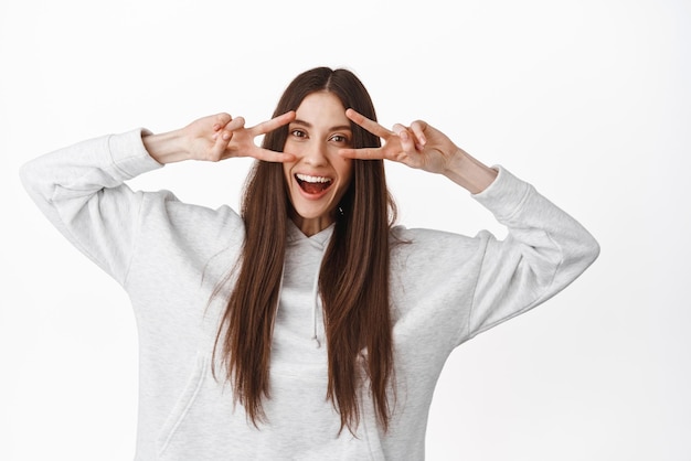 Positive teenage girl with long hair shows peace vsigns and smiles happy at camera dances joyful white background Copy space