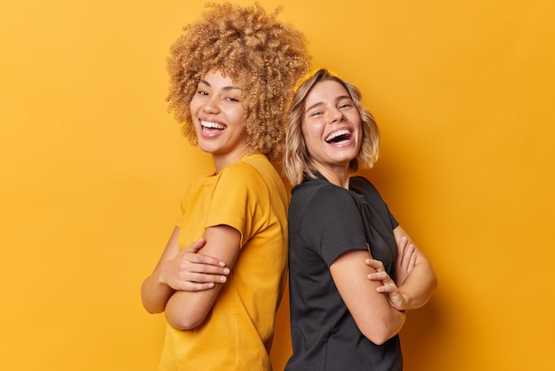 Free photo positive two young pretty women stand back to each other keep arms folded laugh happily dressed in casual t shirts laugh at something funny isolated over yellow background friendship concept