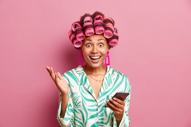 Positive woman dressed in silk slumber suit uses mobile phone downloads new application makes curly hair with rollers isolated over pink wall