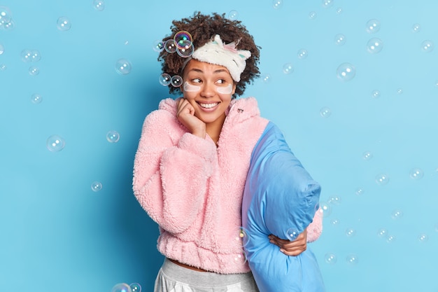 Free photo positive woman enjoys time for rest dressed in warm pajama wears sleepmask on forehead beauty patches poses with pillow against blue wall soap bubbles around