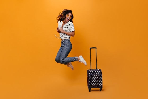 Positive woman in glasses jumps on orange background with tickets for vacation. Joyful adult woman in sunglasses and black polka dot blouse rejoices at camera.