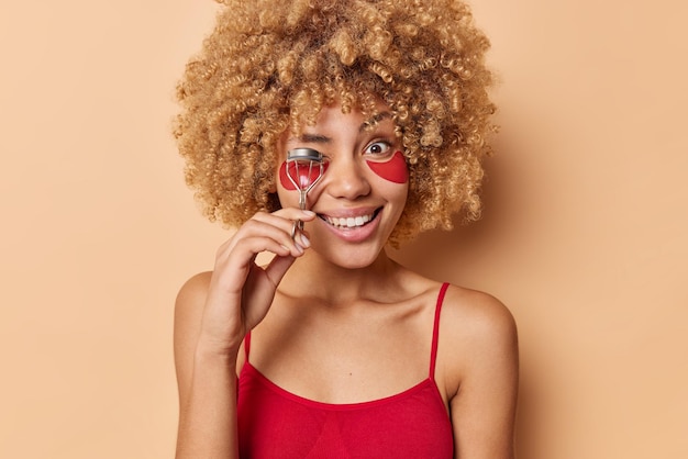 Free photo positive woman has curly bushy hair uses eyelash curler for makeup applies hydrogel patches under eyes dressed in casual red t shirt undergoes beauty treatments isolated over beige backgound