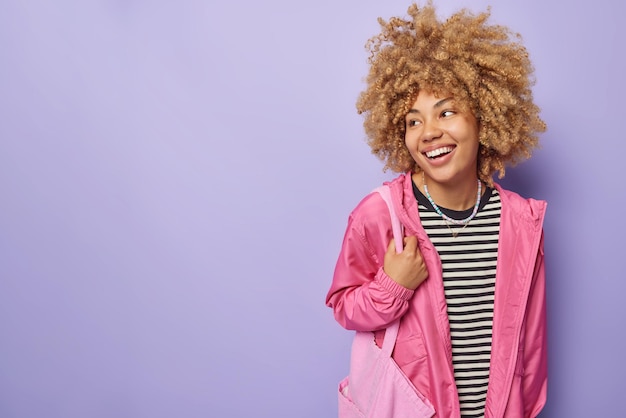 Free photo positive woman has curly bushy hair wears casual striped jumper pink windbreaker carries fabric bag looks away glad to hear good news isolated over purple background blank space for your promo