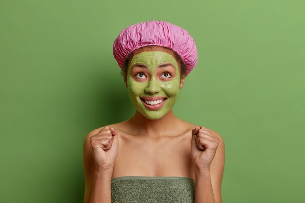 Positive woman looks happily above clenches fists awaits for something special wears bath hat and towel around body applies nourishing avocado mask on face isolated on green wall