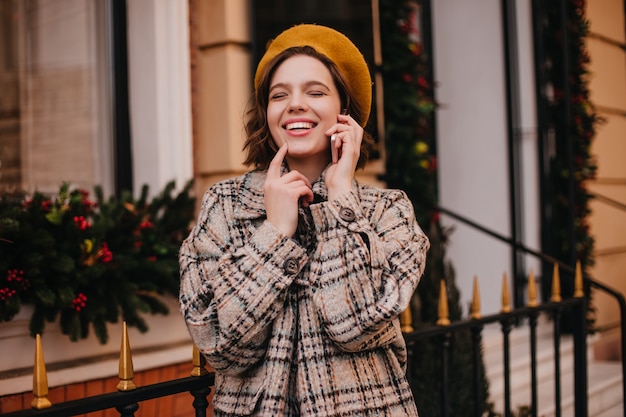 Free photo positive woman in orange beret and coat laughs while talking on phone against city wall