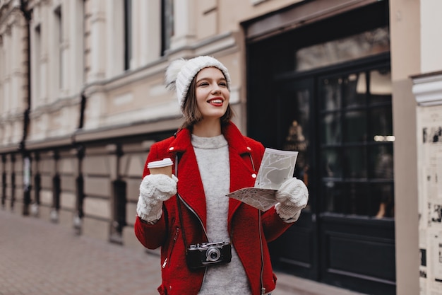 Positive woman in red warm jacket, cashmere sweater and white hat with gloves walks around city with coffee. Tourist with retro camera around her neck holds map.