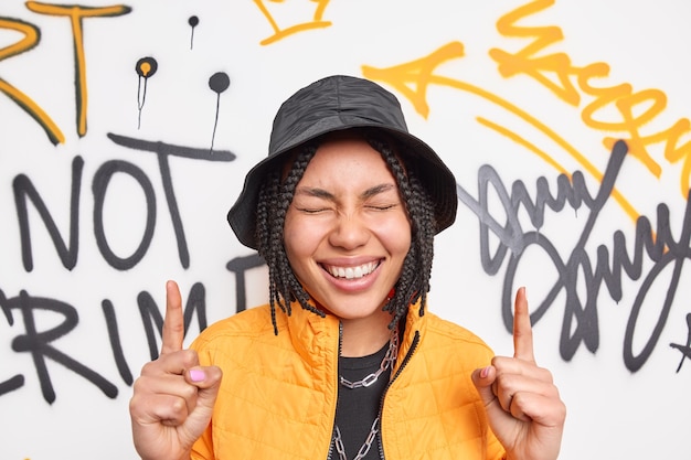 Free photo positive woman smiles broadly points above with two index fingers has happy expression dressed in fashionable clothes poses against graffiti wall