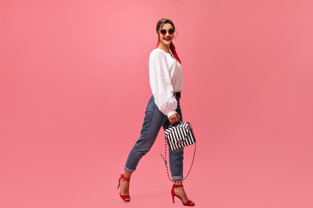 Positive woman in stylish outfit moves on pink background.  Pretty woman in white blouse and red high heels is smiling at camera.