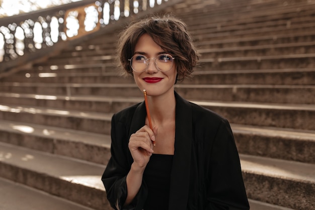 Free photo positive woman with short hair and bright lips in glasses smiling outside. trendy lady in black clothes posing on stairs.