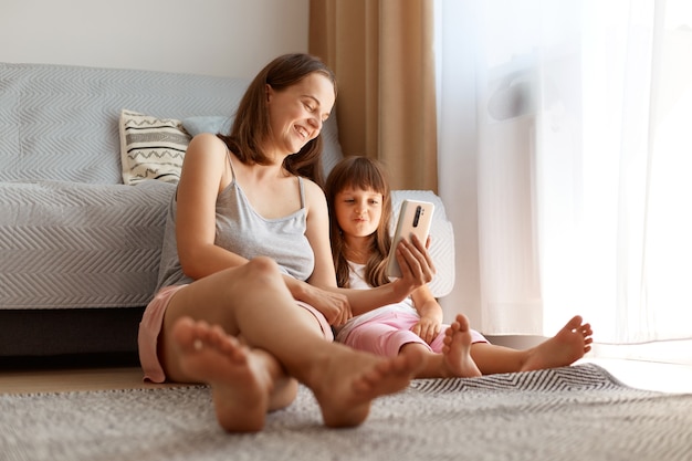 Positive young adult Caucasian female wearing casual style clothing posing indoor in living room with her daughter while sitting on the floor near sofa and window, watching funny video with child.