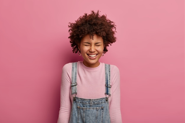 Free Photo positive young afro american girl smiles broadly, has upbeat mood, laughs at something very funny or hilarious, closes eyes, hears crazy joke, dressed casually, stands against pastel rosy wall